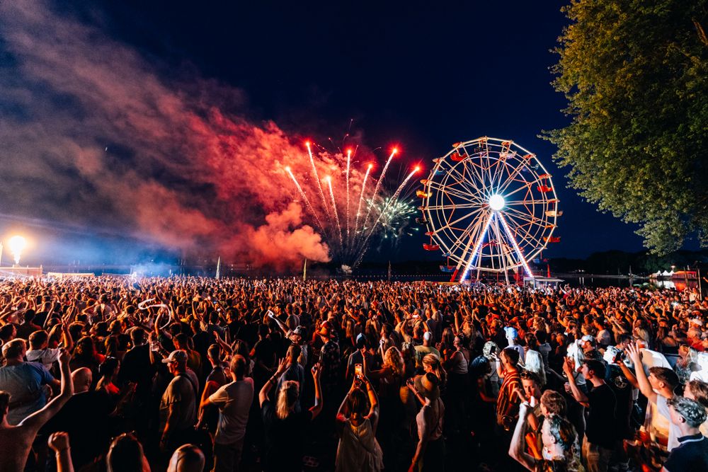 Nighttime festival crowd watching fireworks display next to illuminated Ferris wheel.