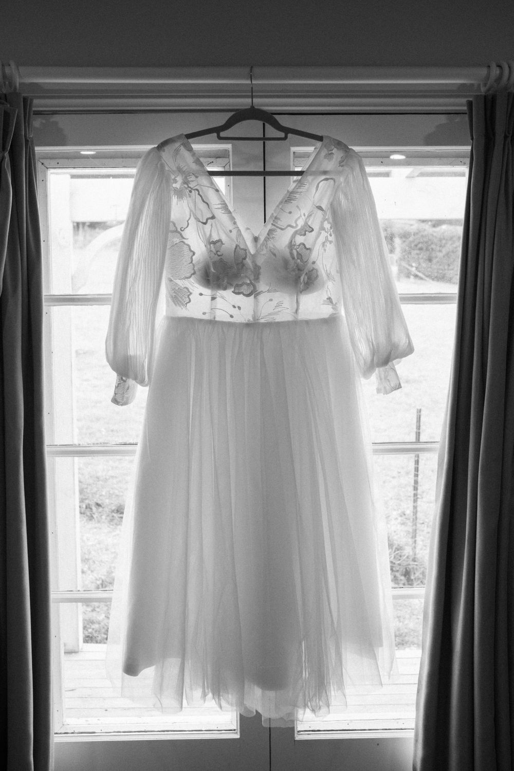 Elegant white wedding dress with lace bodice and sheer sleeves hanging in window light.