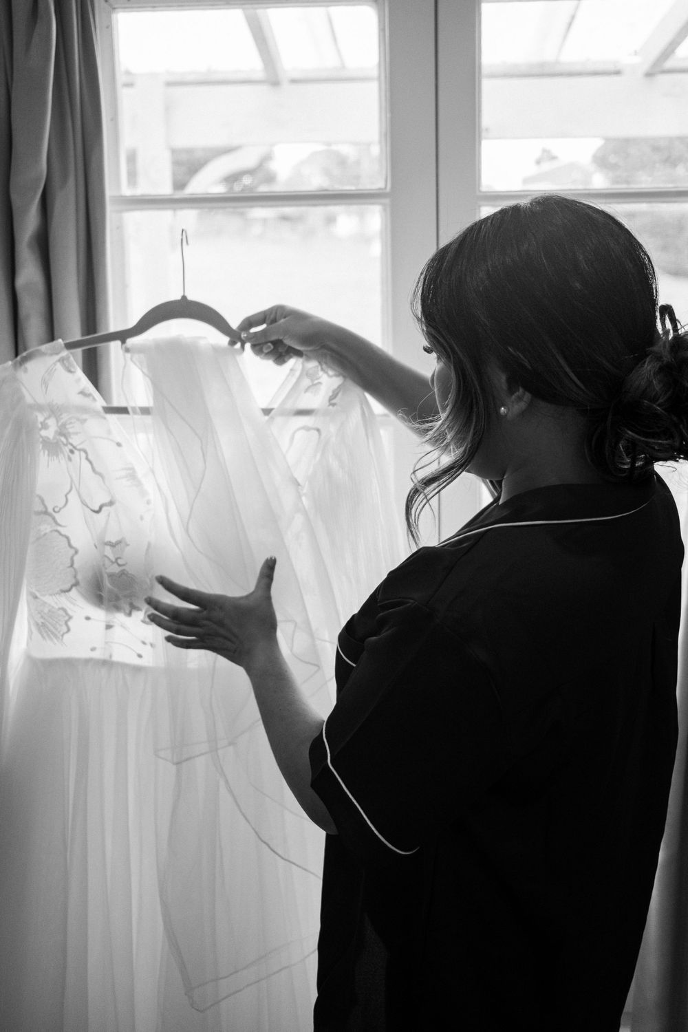 Silhouette examining a white wedding dress hanging by a sunlit window.