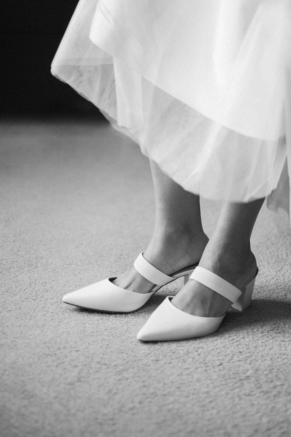Close-up view of white bridal shoes with delicate straps peeking out from beneath a flowing white dress.