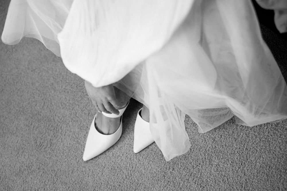 Black and white detail shot of white bridal shoes beneath flowing wedding dress fabric.