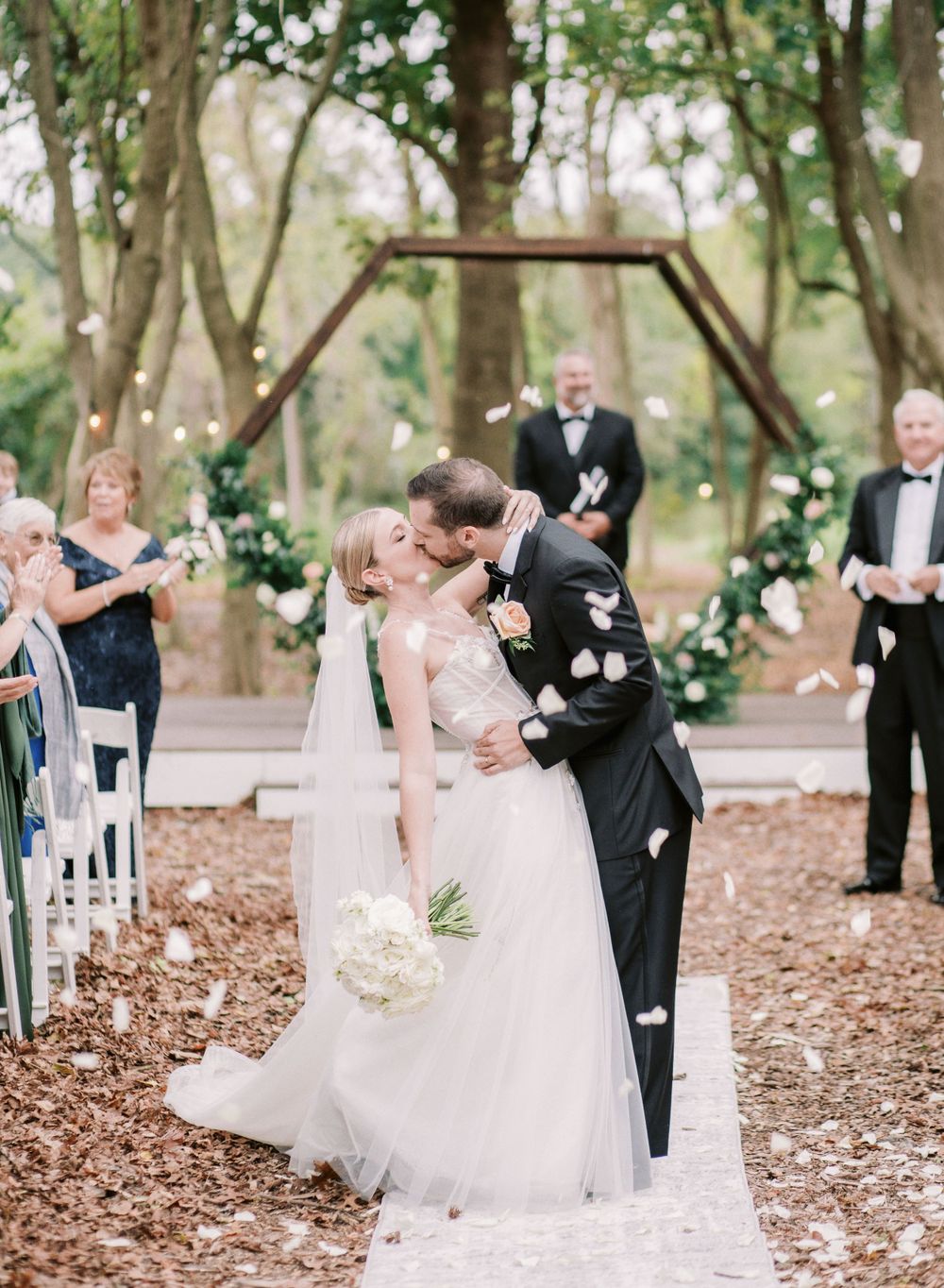 Outdoor forest wedding ceremony with flower petals falling during a special moment under wooden arch.