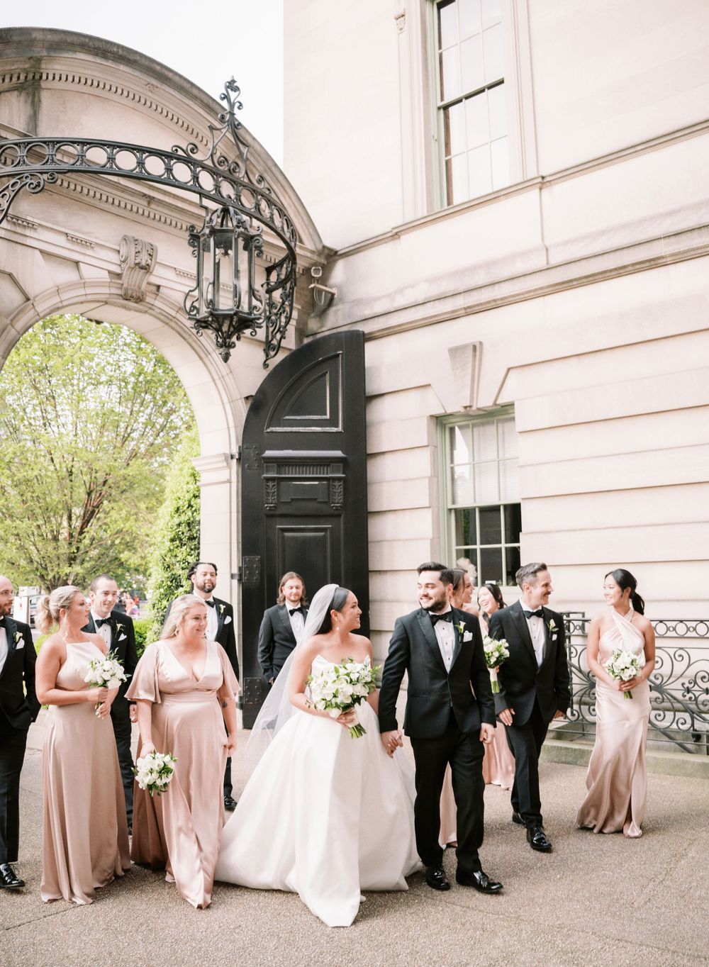 Elegant wedding party in formal attire gathered outside historic building with ornate architecture.