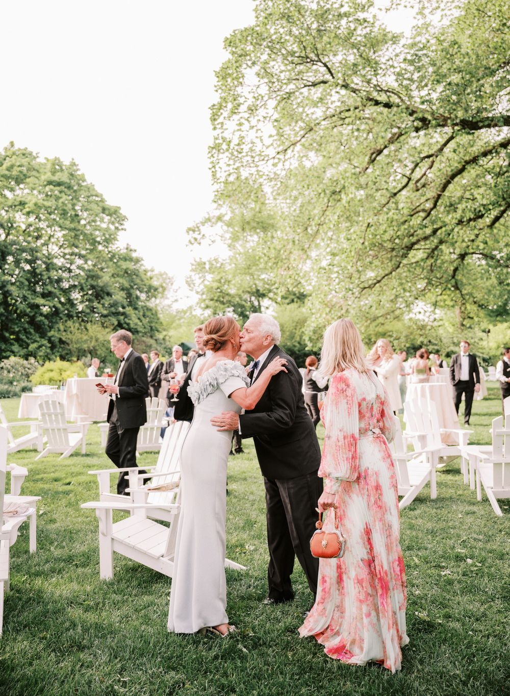 An outdoor garden wedding ceremony with guests gathering among white chairs under leafy trees.