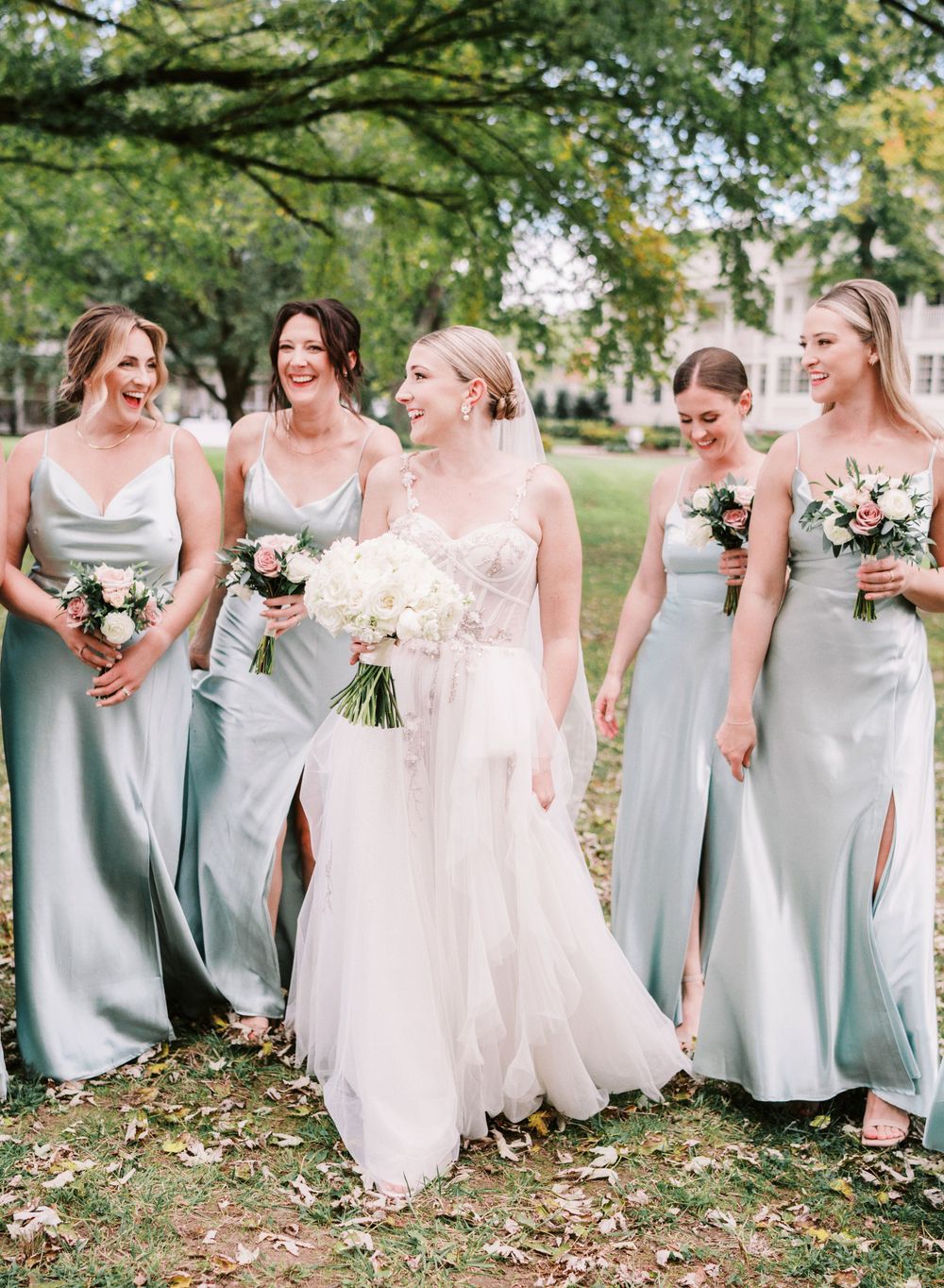 Wedding party in sage green bridesmaid dresses walking and laughing together outdoors.