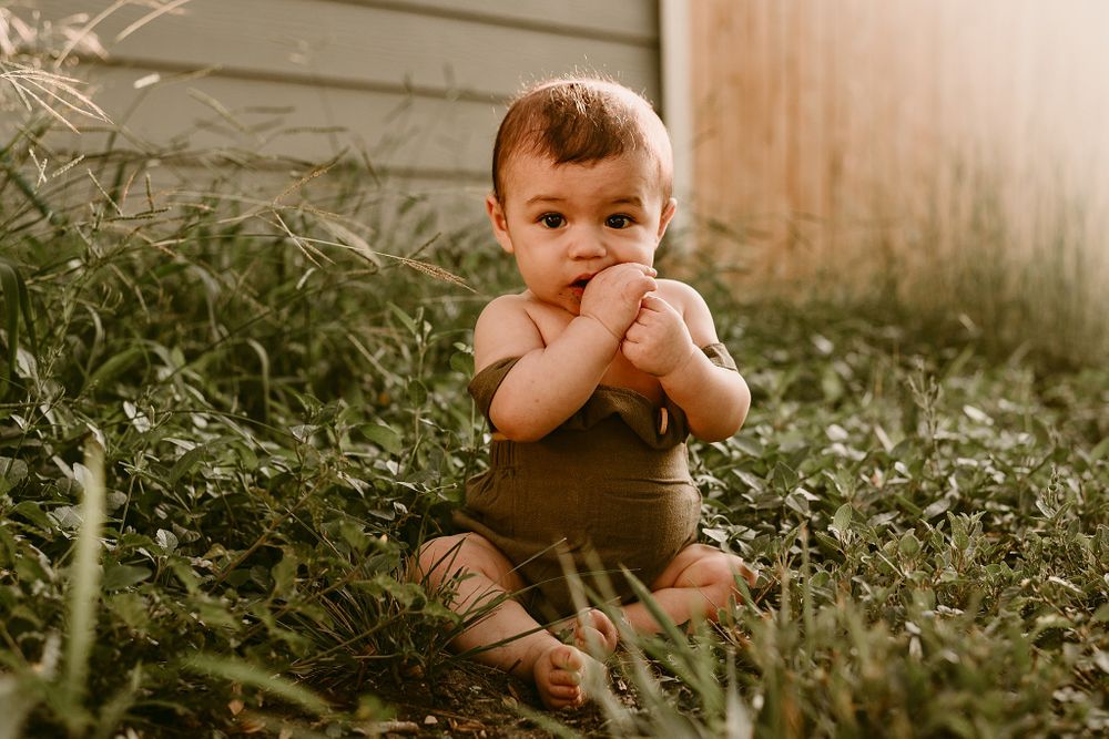 Toddler in brown outfit sitting among garden plants with hands near face.