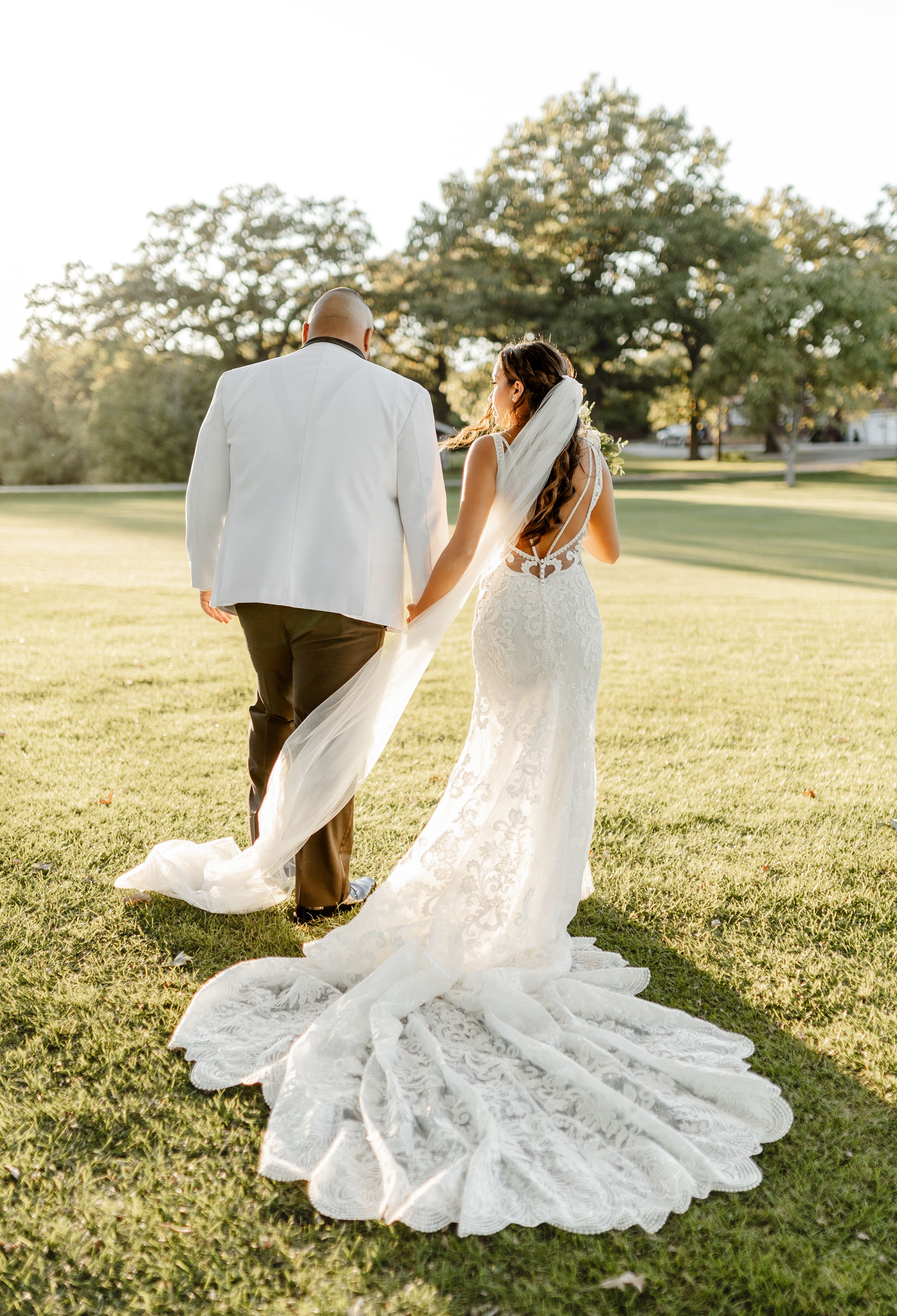 A couple walks hand in hand across a grassy lawn with the bride's detailed lace train flowing behind.