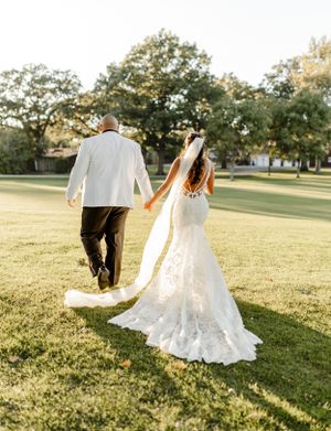 Couple strolls across a sunlit lawn with the bride's flowing lace gown trailing behind them.