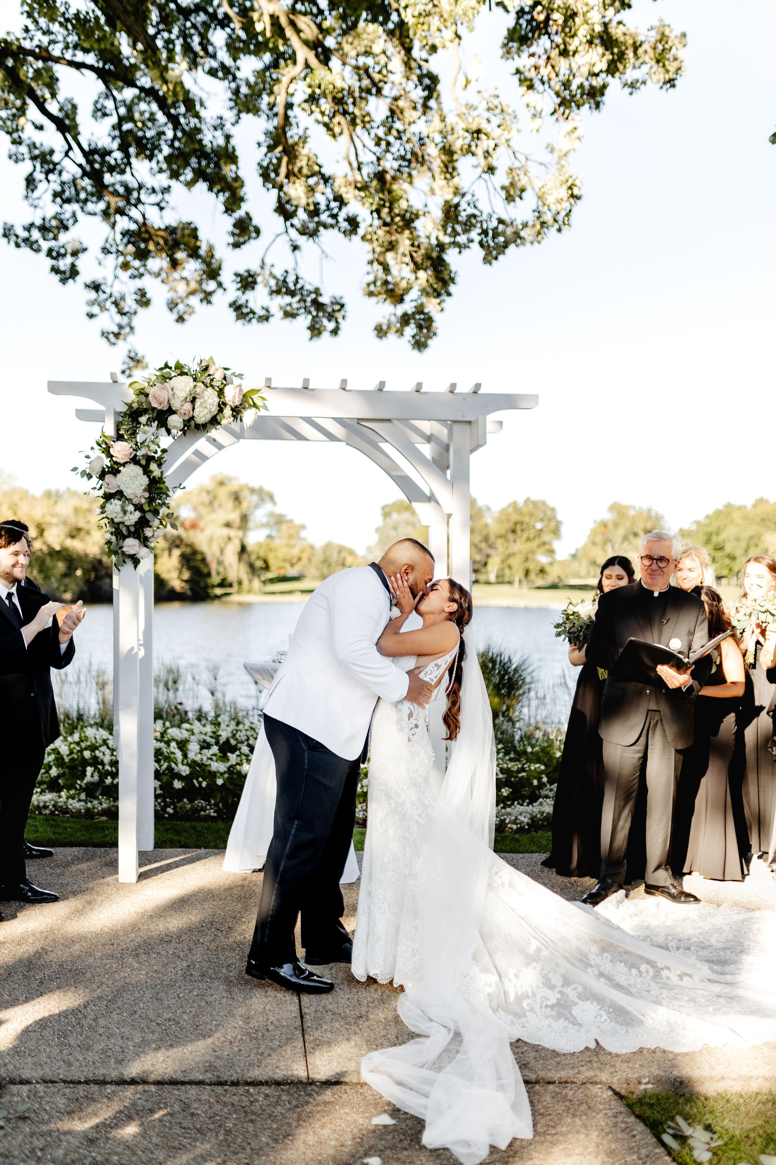 Wedding ceremony under a white pergola by a lake with guests witnessing the couple's first kiss.