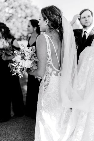 Black and white photo capturing bride in detailed lace gown holding bouquet during ceremony.