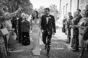 Wedding guests throw confetti over a couple walking down an outdoor brick pathway.