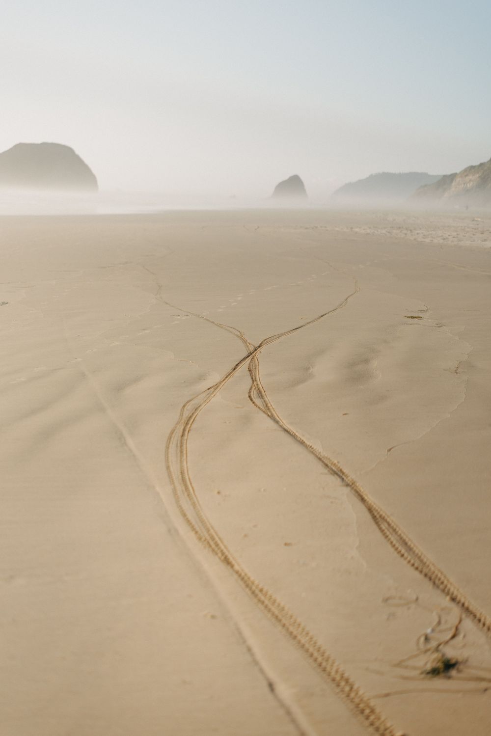 Tire tracks intersect on misty beach with coastal rock formations in distance.