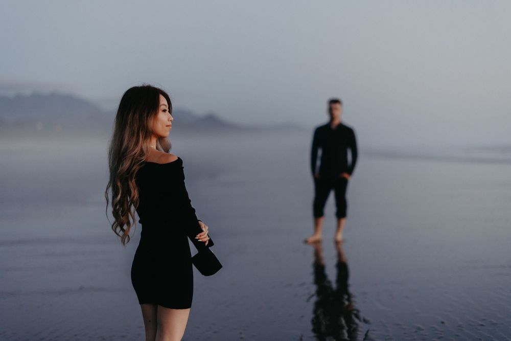 Moody beach scene with two people in dark clothing against misty coastal backdrop.