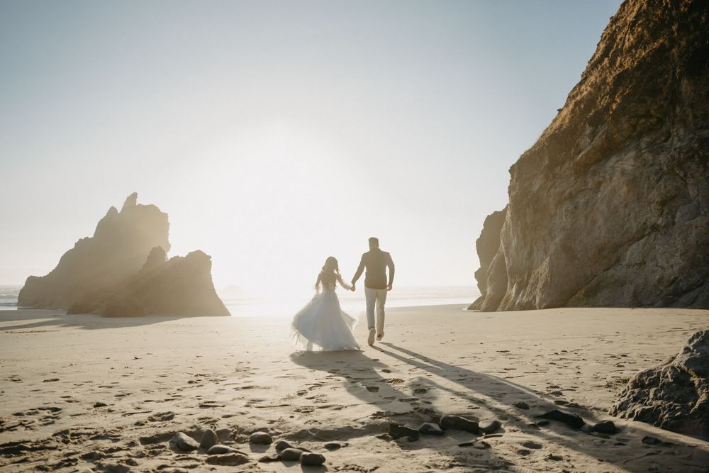 A couple in wedding attire walking on a beach at sunset with dramatic rocky cliffs in the background.