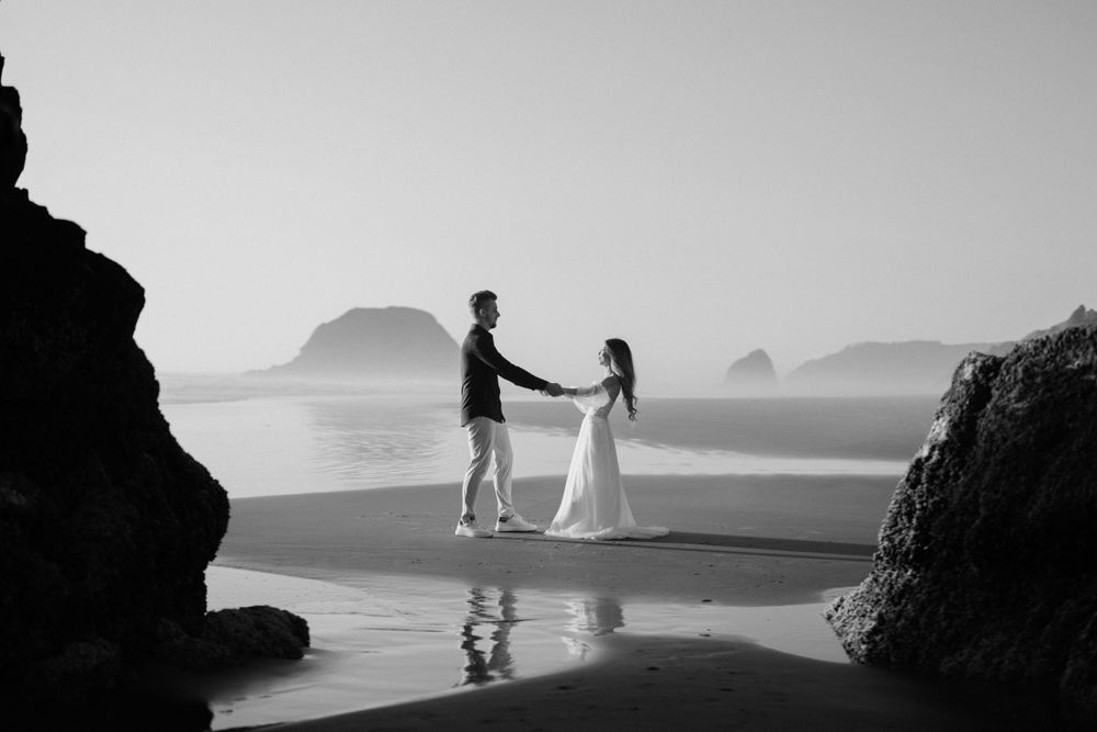 Black and white beach wedding photo with couple standing between rock formations.
