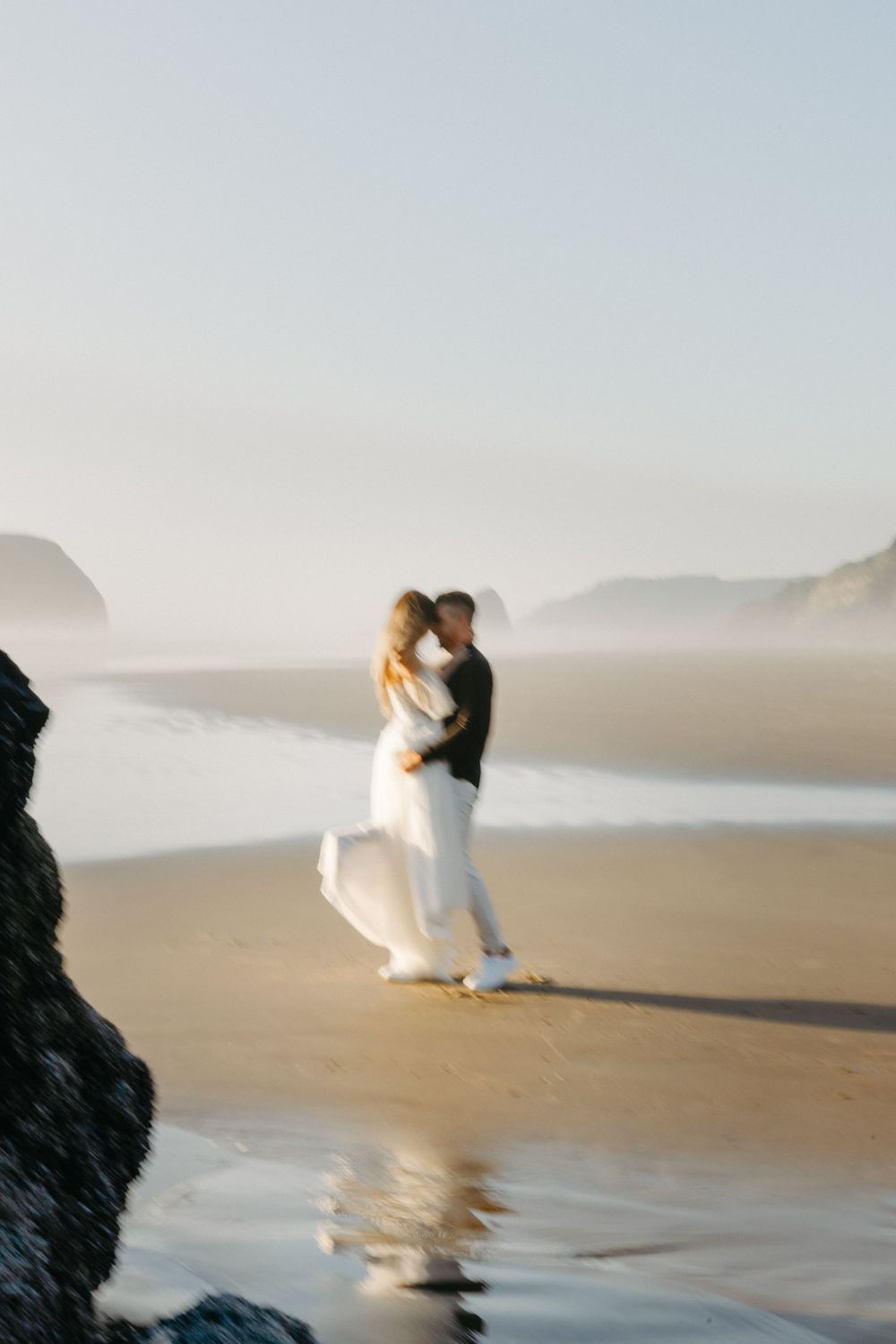 A romantic couple embraces on a misty beach with waves crashing in the background.