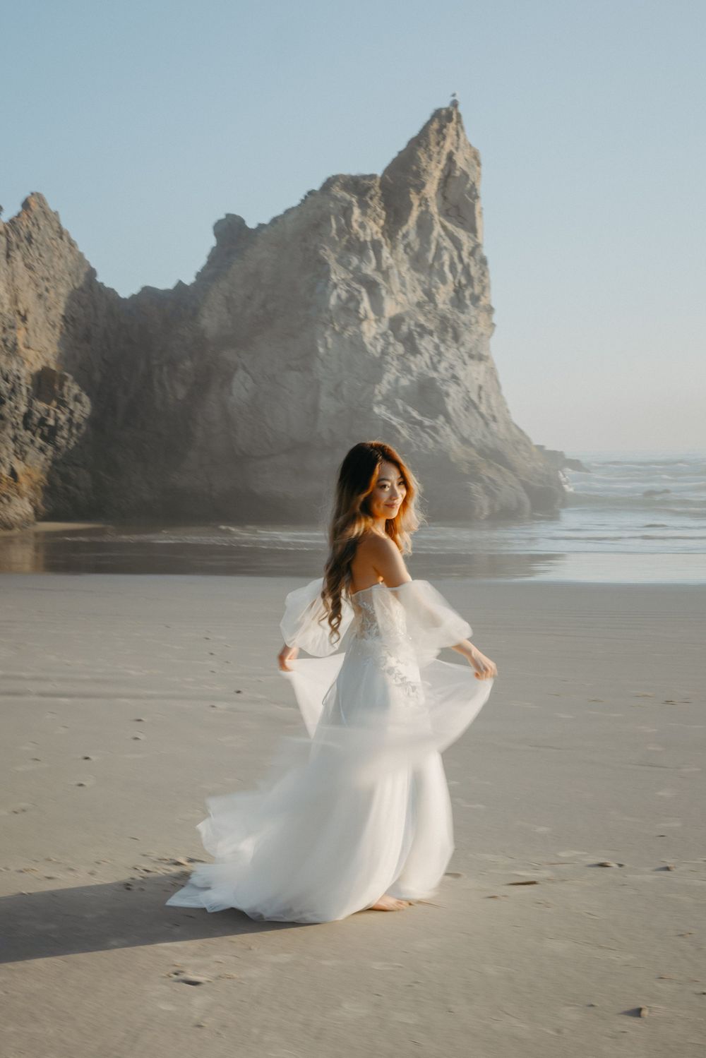 A flowing white wedding dress twirls on a beach with dramatic coastal rock formations in the background.