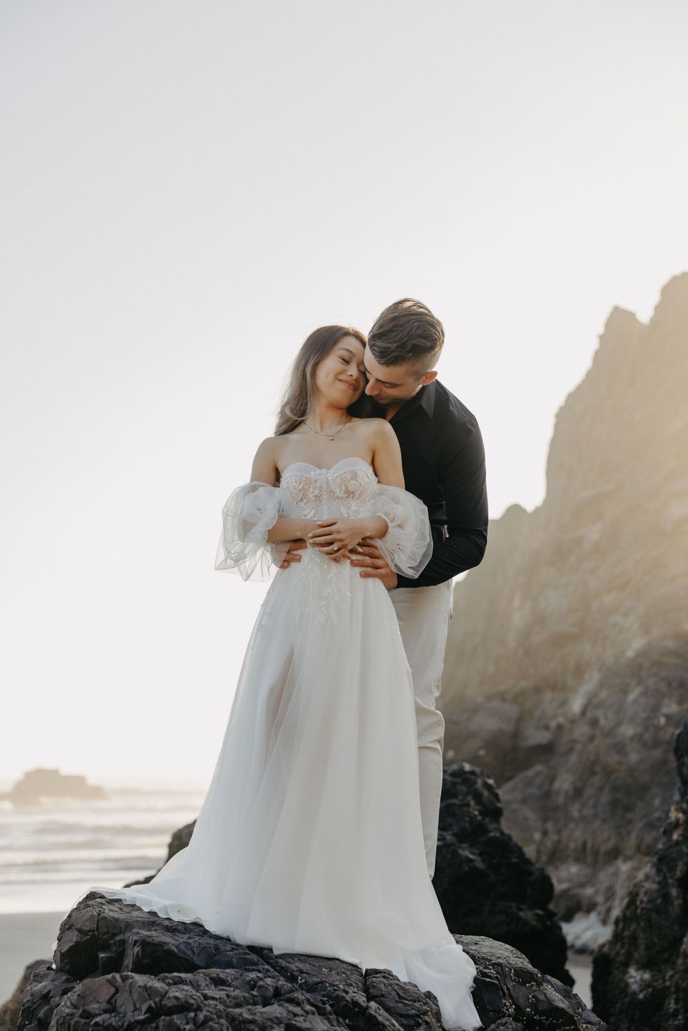 A romantic moment between a couple in wedding attire standing on coastal rocks at sunset.