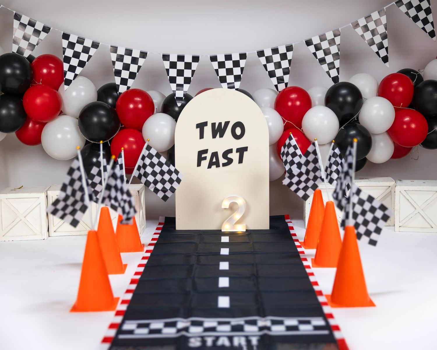 Racing themed party backdrop with checkered flags, red and black balloons, and orange traffic cones around a black runway.