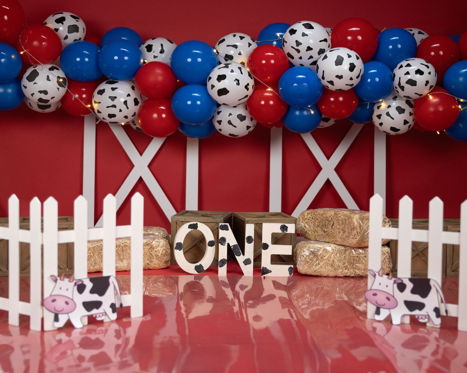 Barn-themed birthday display with 'ONE' sign, white picket fence, and red, blue and cow print balloons.