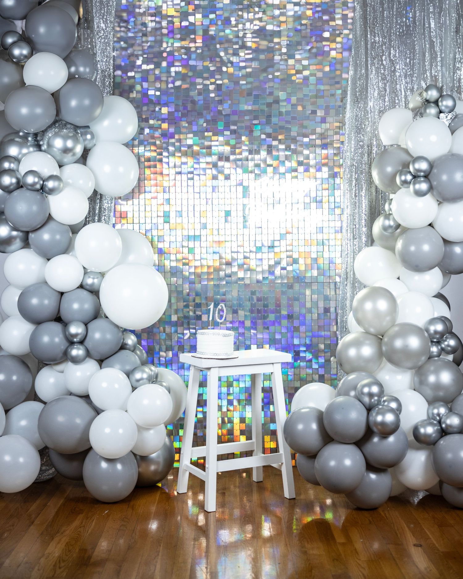 Modern party backdrop with gray and white balloon arch against holographic wall and white stool.