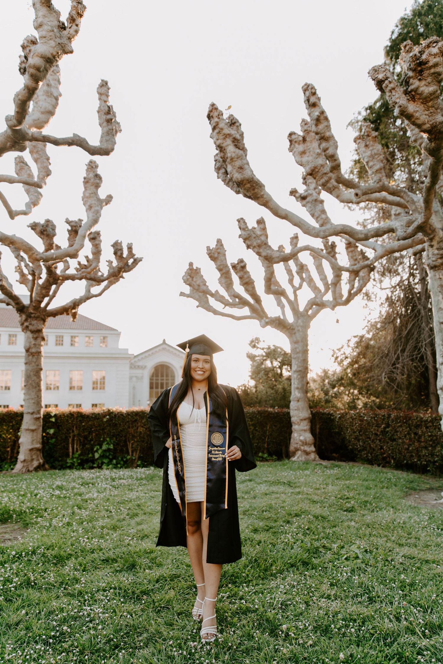 Graduate poses in academic regalia among bare winter trees on campus lawn.