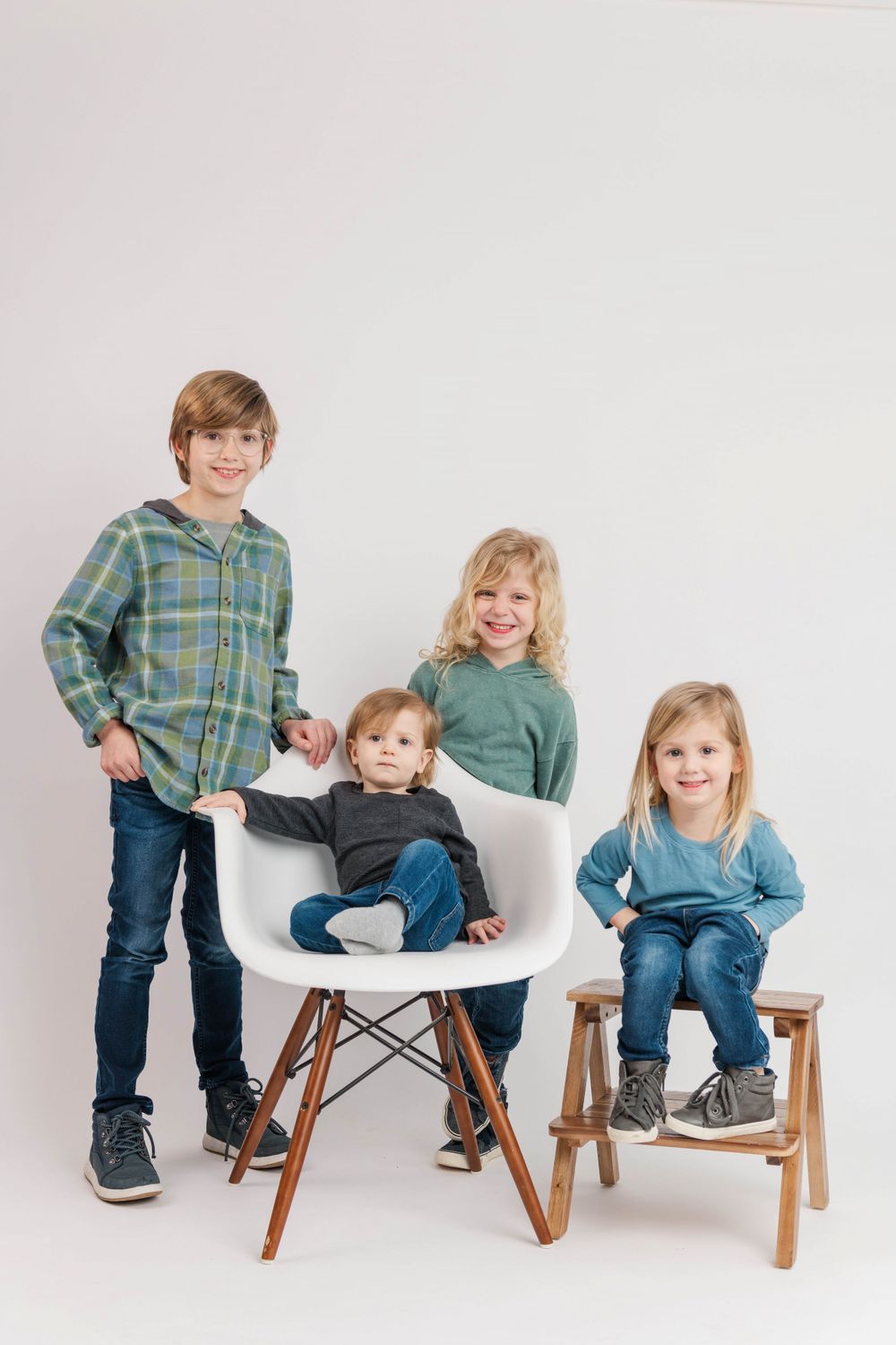 Four children in casual clothing sitting and standing around white chairs against a neutral background.