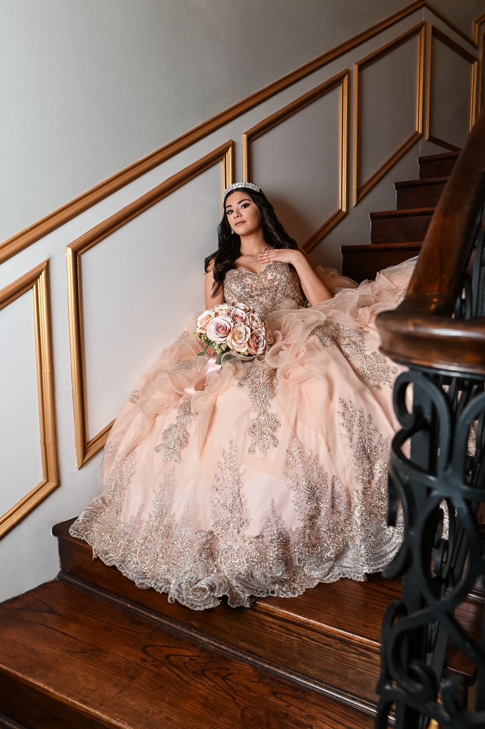 Elaborate quinceañera dress with layered tulle and lace details on wooden staircase with gold trim.