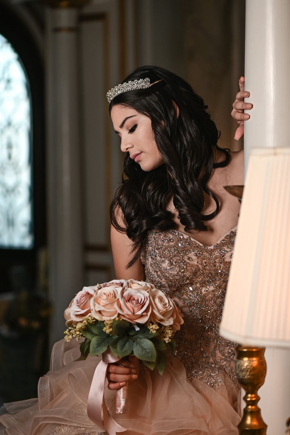 A young woman in a blush pink quinceañera gown holds a bouquet of roses while posing against an elegant pillar.