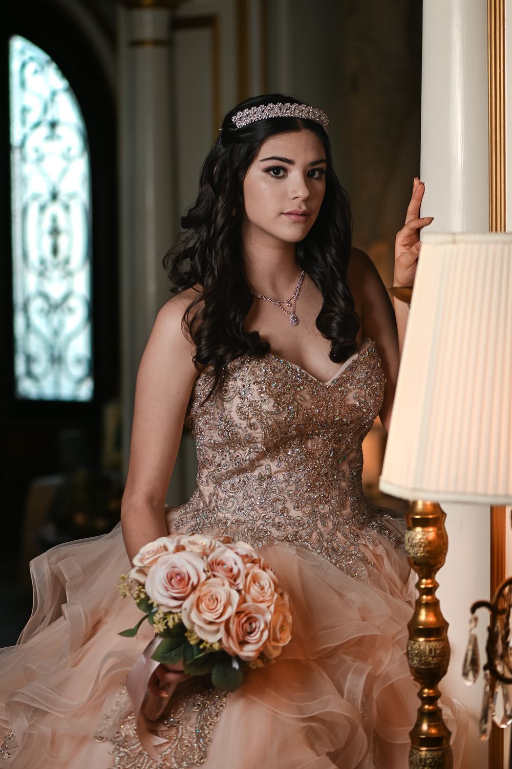 An elegant portrait featuring a crystal tiara and embellished pink ballgown with a bouquet of roses.