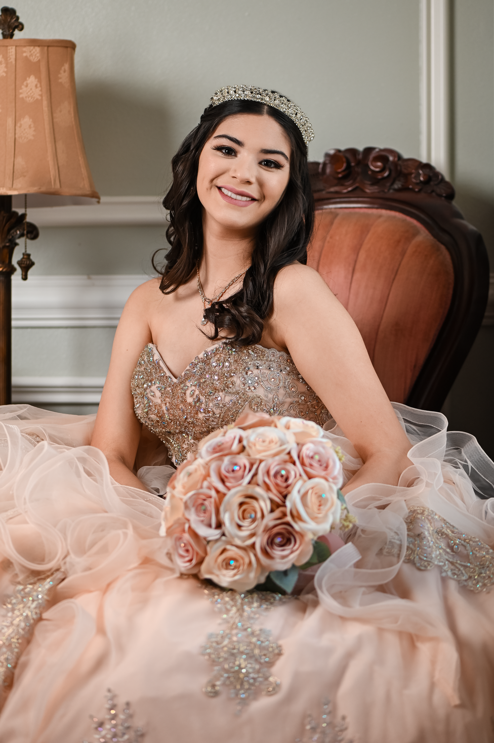 A joyful portrait showcasing a sparkling tiara and blush quinceañera gown with rose bouquet.