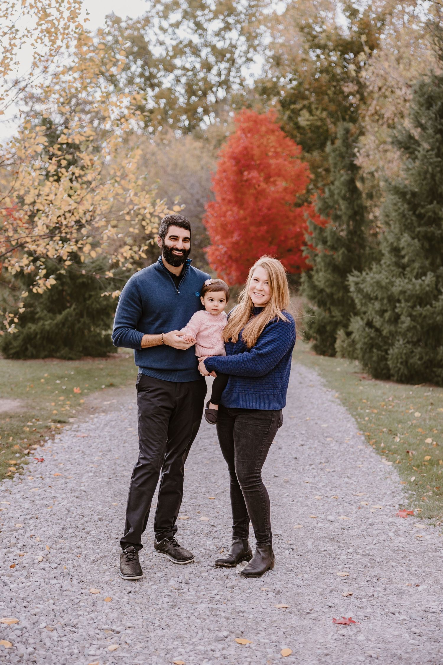A family of three poses together on a path with colorful fall foliage in the background.