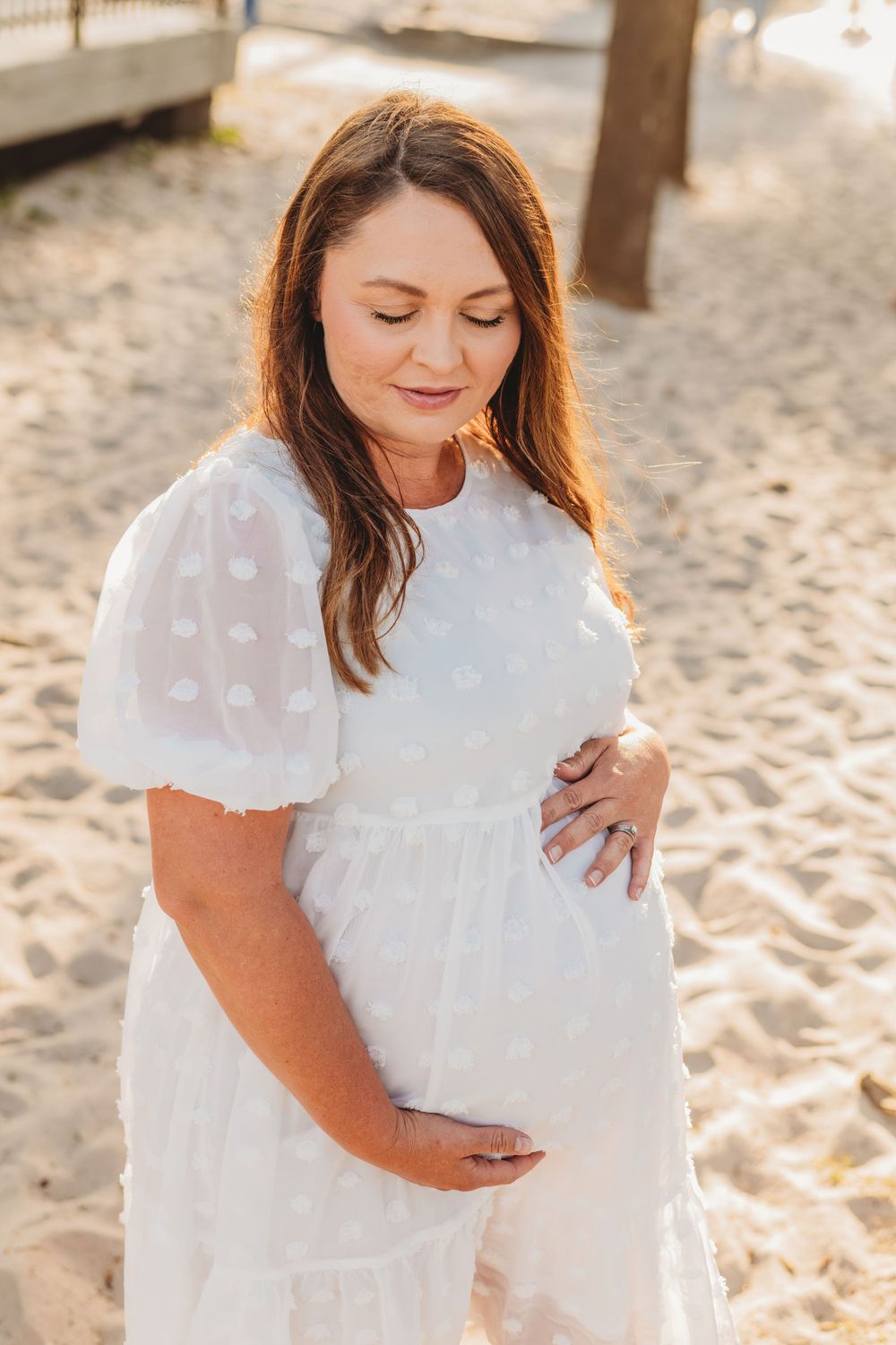 Woman in white dress cradles her baby bump on sunny beach.