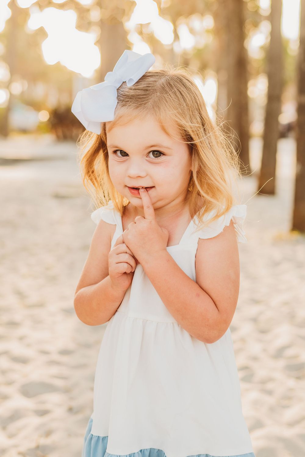 Young child in white dress plays peek-a-boo on sandy beach with palm trees in background during golden hour.