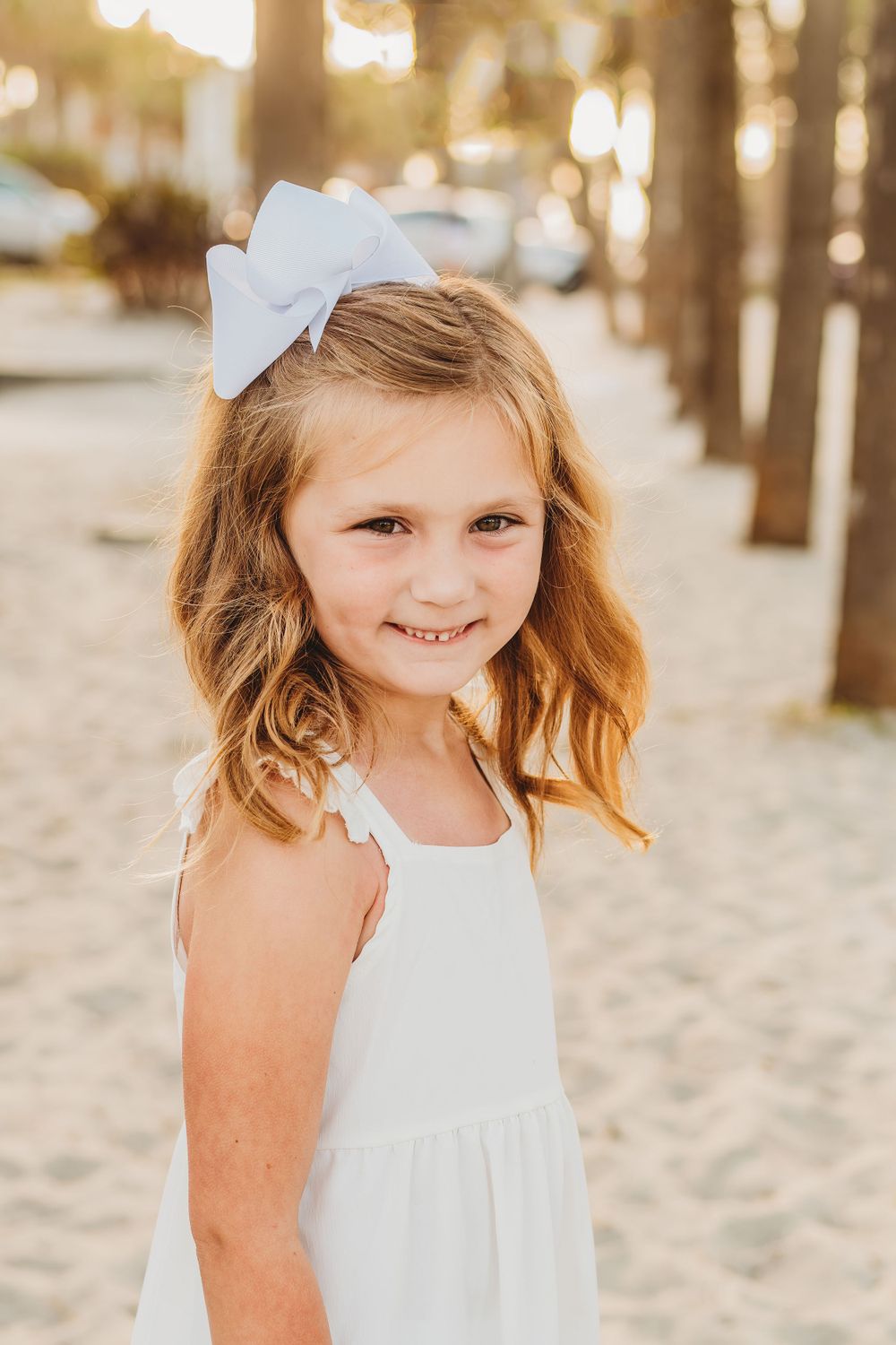 Young girl in a white dress smiling on a sandy beach path at sunset.