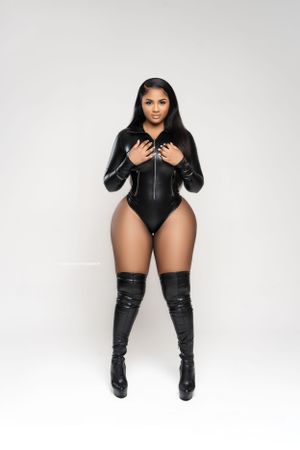 Model in black leather bodysuit and thigh-high boots posing against white background.