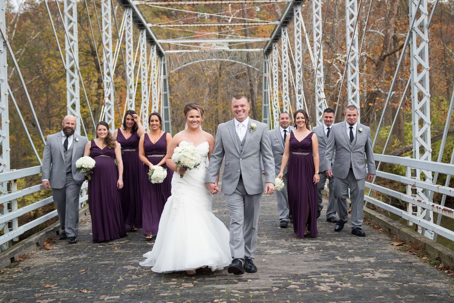 Wedding party poses on a rustic bridge with bridesmaids in purple dresses and groomsmen in gray suits.