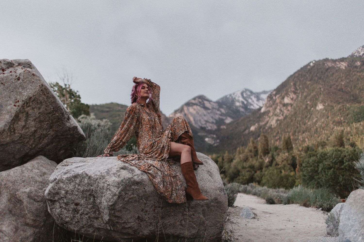 Person in bohemian dress sitting on large boulder with mountain landscape backdrop.