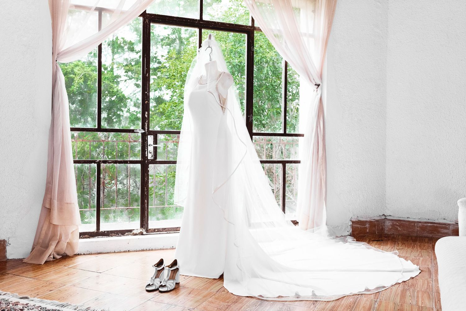 Elegant white wedding dress hanging in sunlit dressing room with bridal shoes on wooden floor.