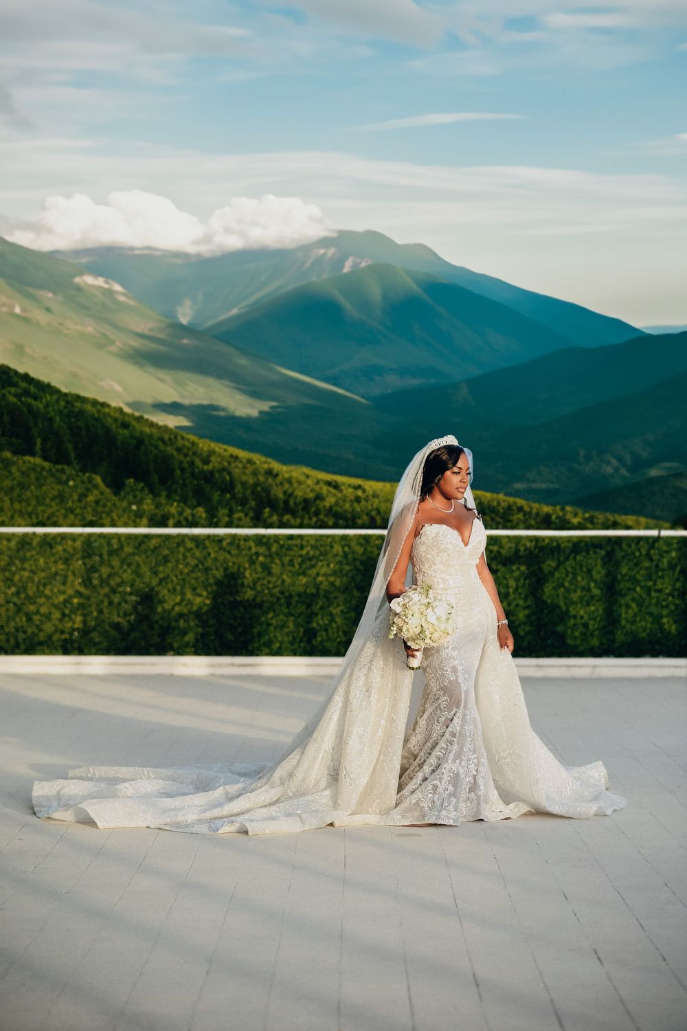 Elegant bride in lace mermaid gown with long veil stands on terrace with stunning mountain landscape backdrop.