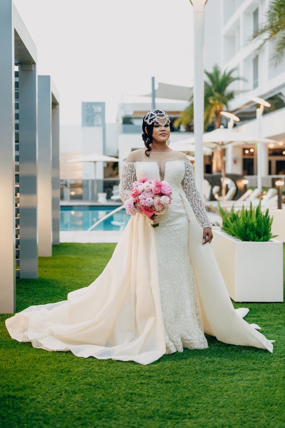 Bride in off-shoulder lace wedding gown with train holds pink bouquet outdoors near pool.