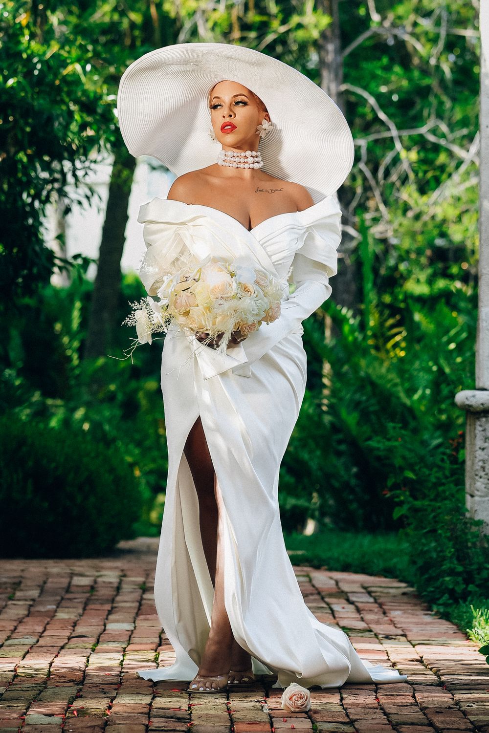 Glamorous bride in dramatic off-shoulder white gown and oversized hat holds bouquet in lush garden setting.