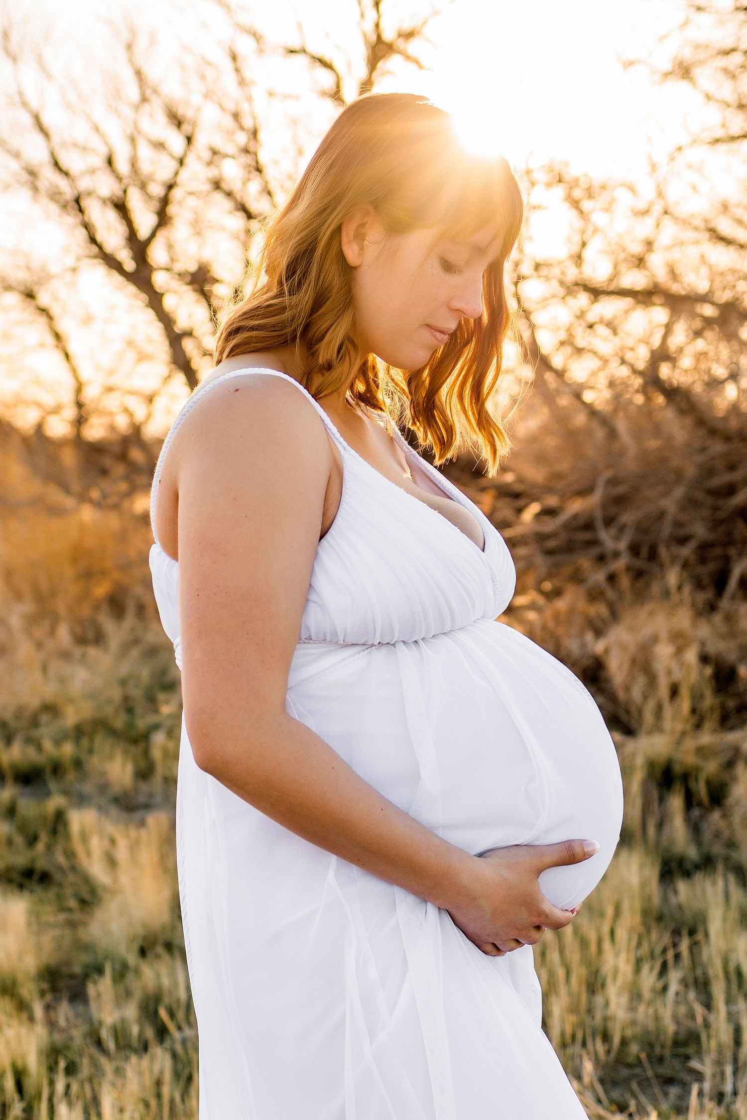 Pregnant woman in long white dress, she is holding her bump, looking down, sun flares over her head