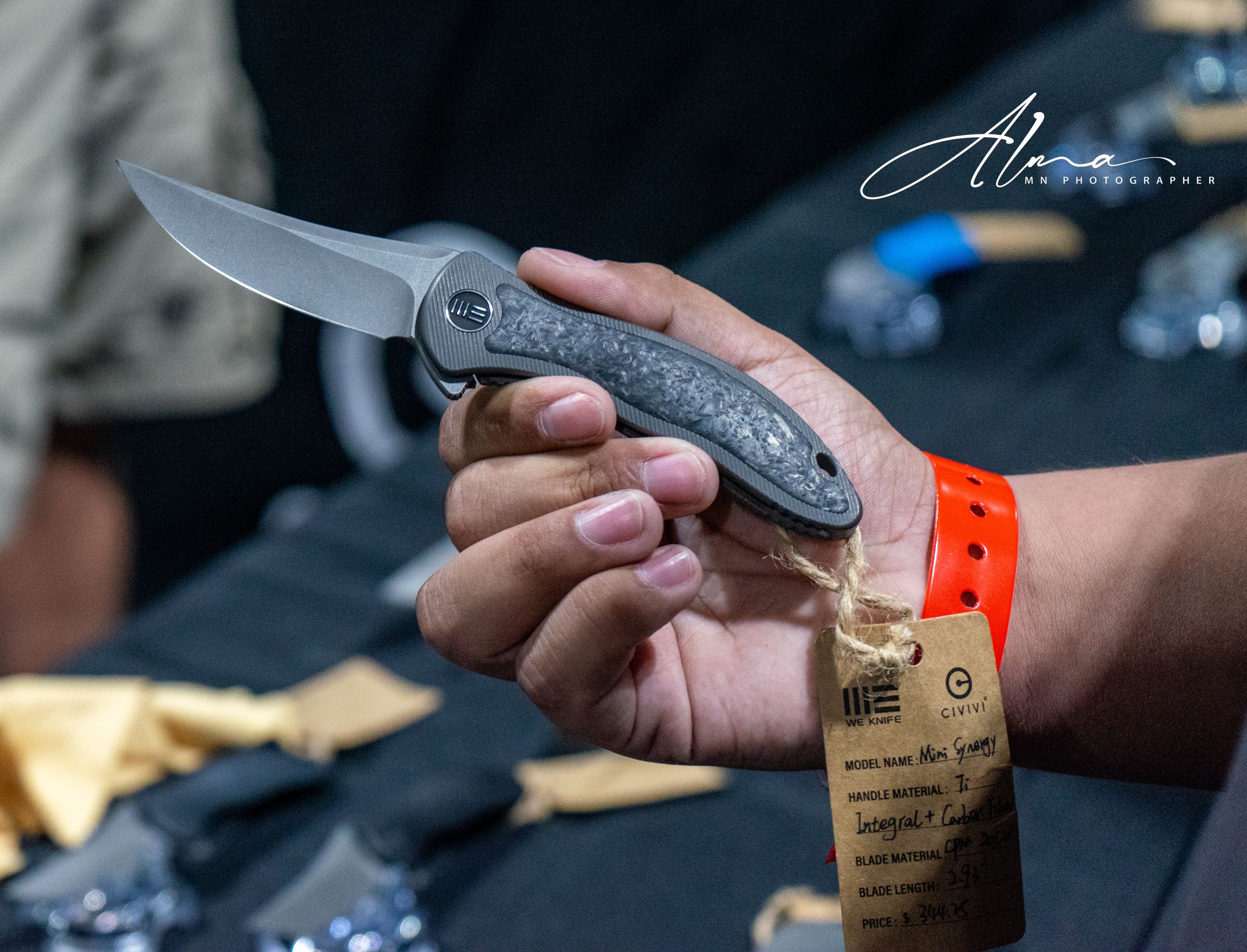 Close-up of hands holding a folding knife against a dark background with an orange wristband visible.