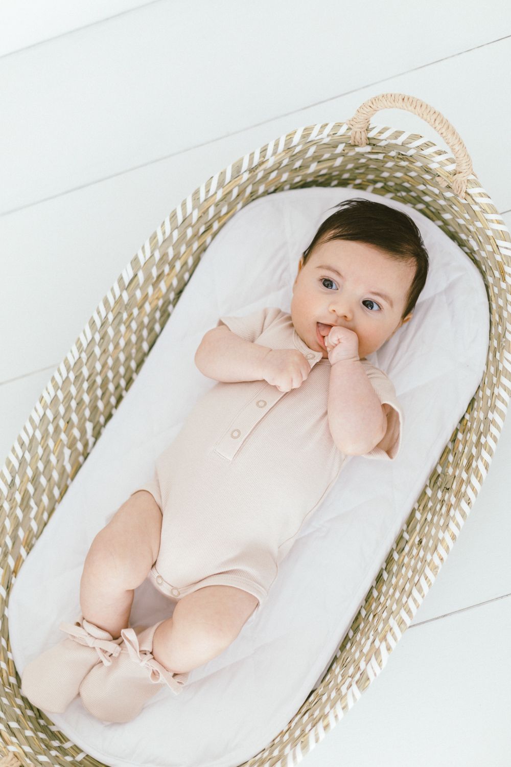 Baby wearing light pink romper rests in oval woven bassinet.