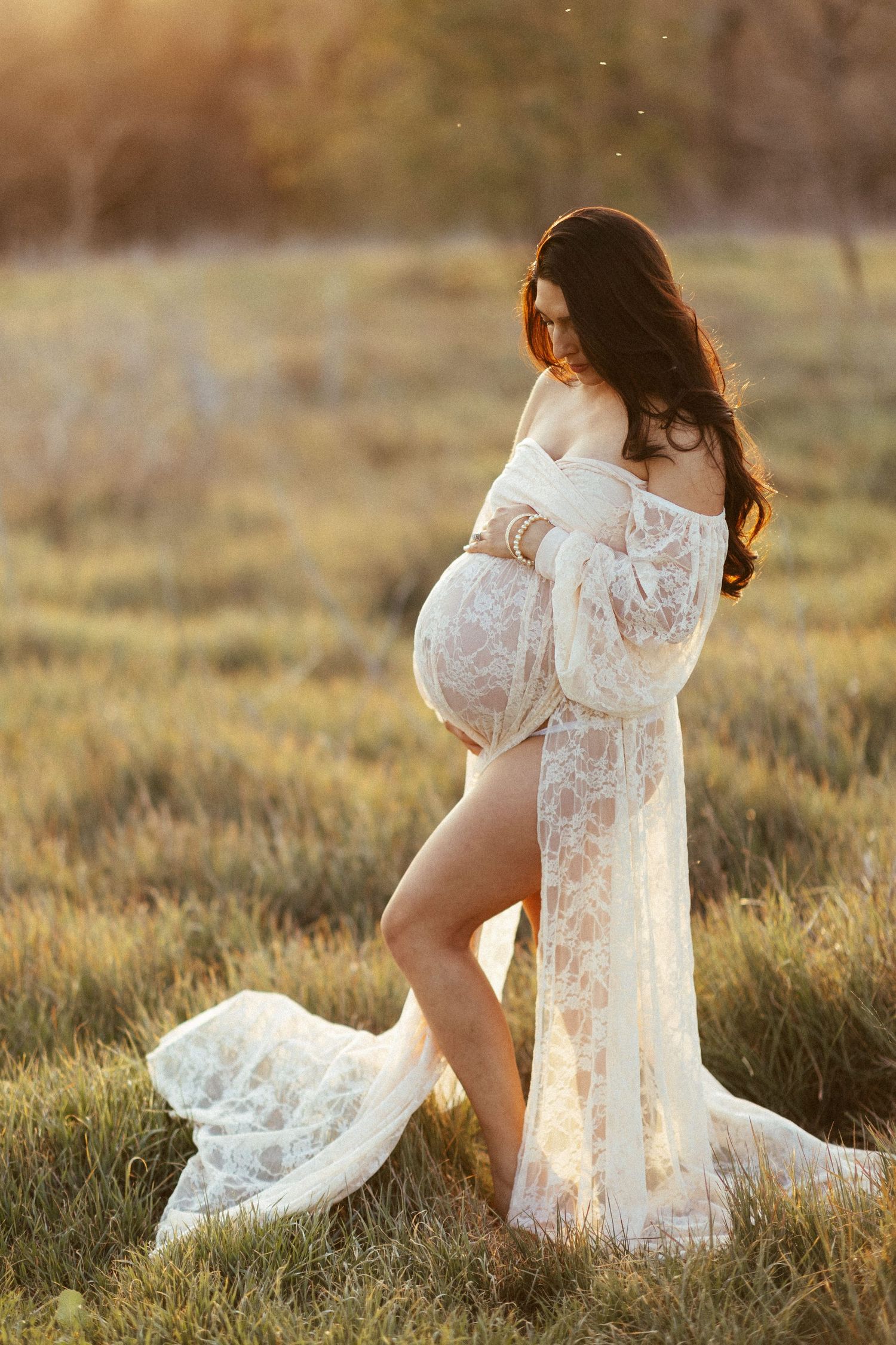 A maternity photo of a woman in a flowing white lace dress at sunset in a field.