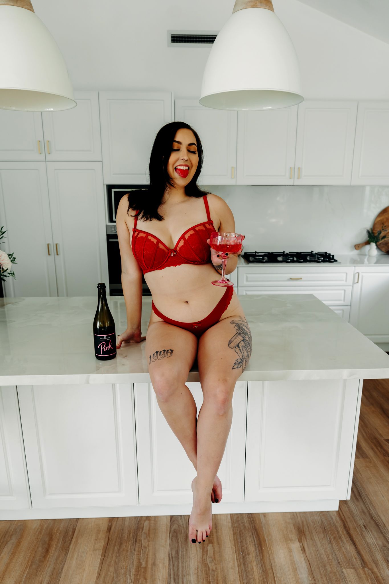 Woman in red lingerie sits on white kitchen counter with cocktail and wine bottle.
