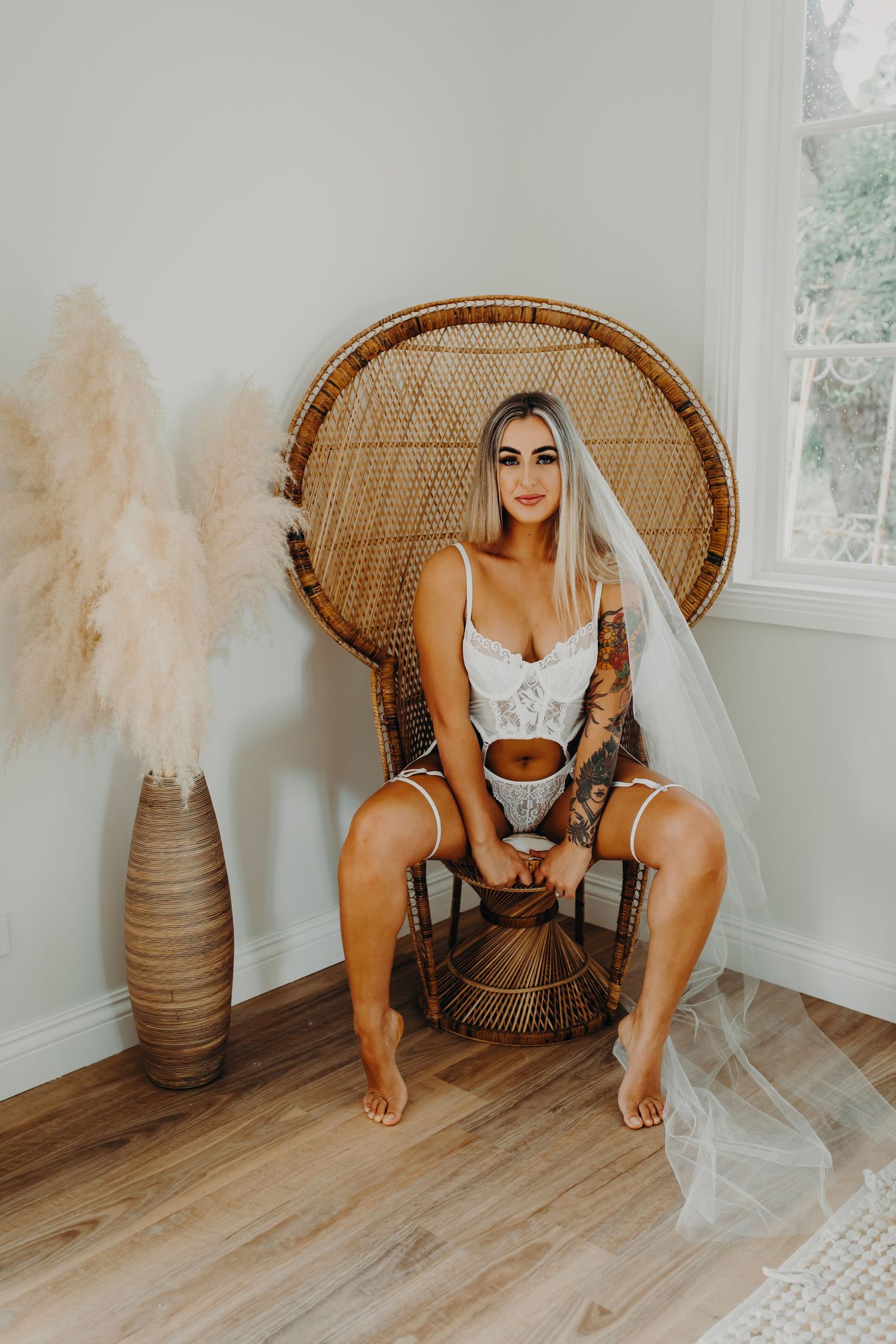 Person in white bridal lingerie sitting on rattan peacock chair with tattoos and veil.