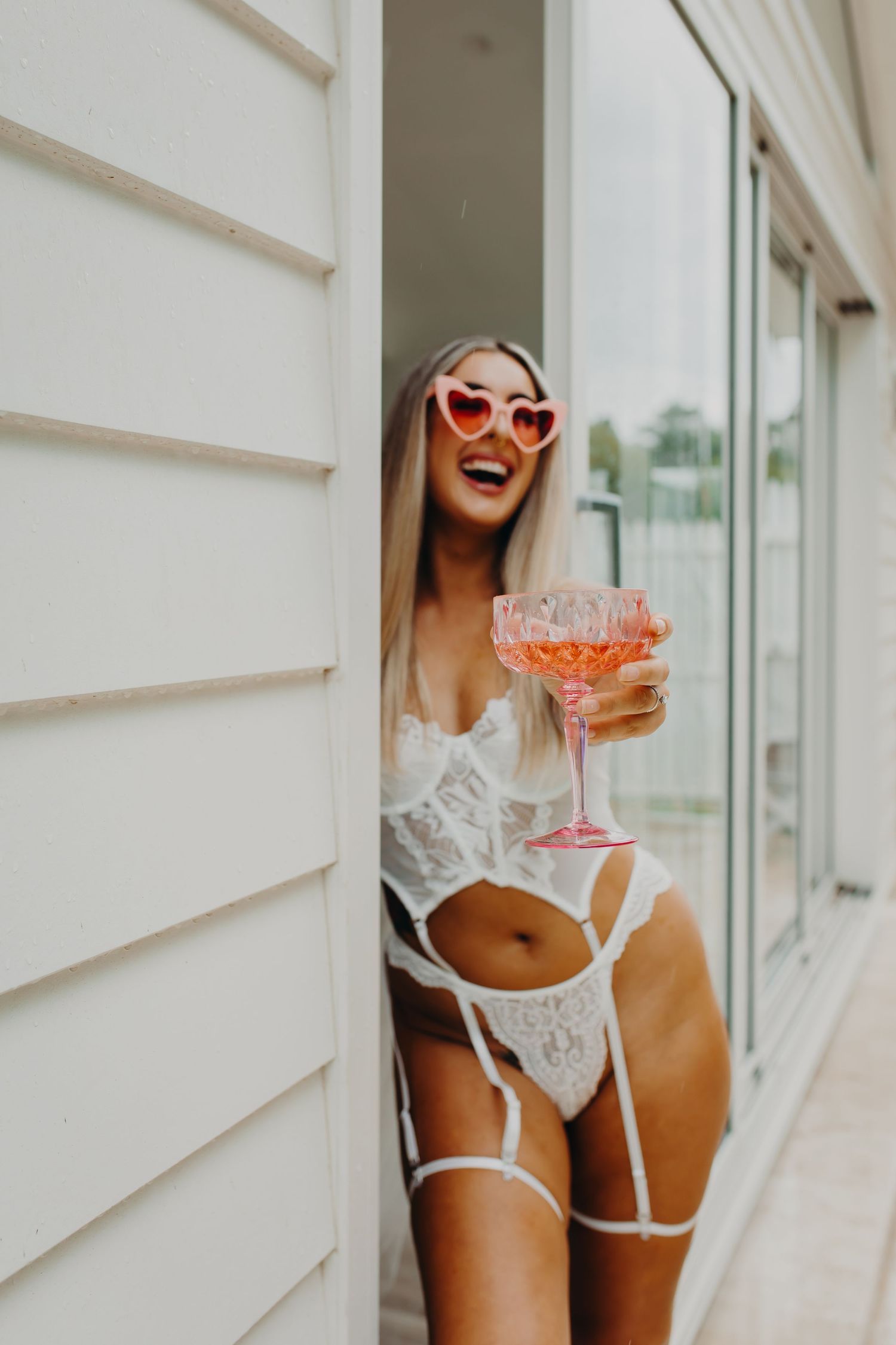 Person in white lingerie holding pink cocktail while wearing heart-shaped sunglasses.