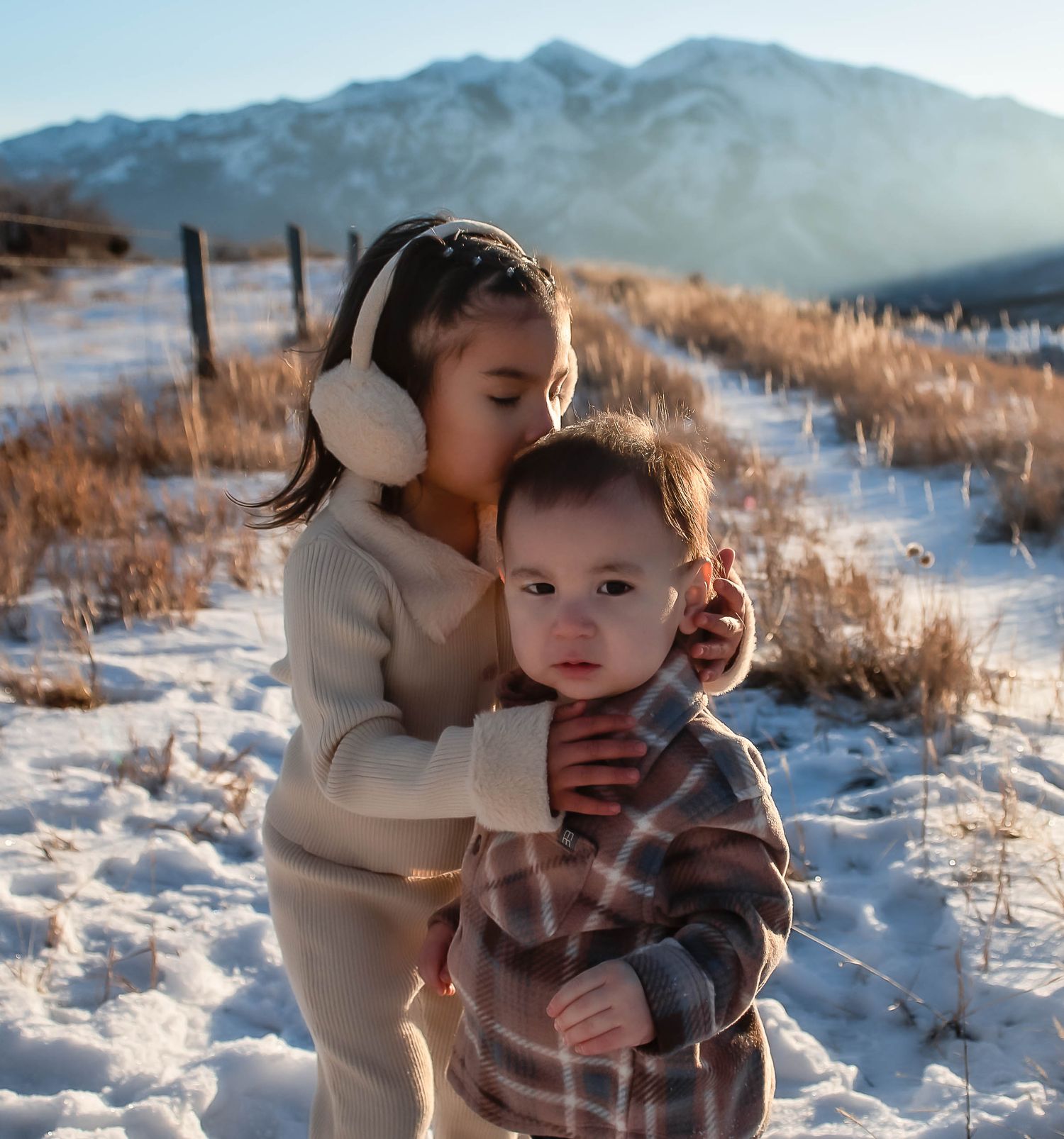 kids on a snowy field with mountain in the background, embracing each other and little sister giving a kiss on brothers head